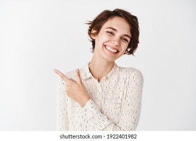 Young caucasian woman with natural beauty without makeup, smiling white teeth and pointing finger left at logo, standing over studio background.
