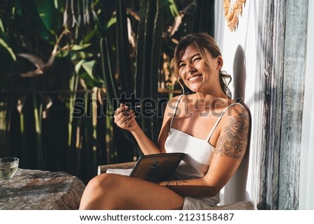 Young caucasian woman with notebook look at camera on terrace of resort hotel. Concept of tourism, vacation and weekend. Pretty smiling girl. Sunny day. Idyllic and tranquil lifestyle on Bali island