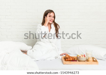 Young caucasian woman on the bed blink at the camera through fingers, embarrassed covering face.