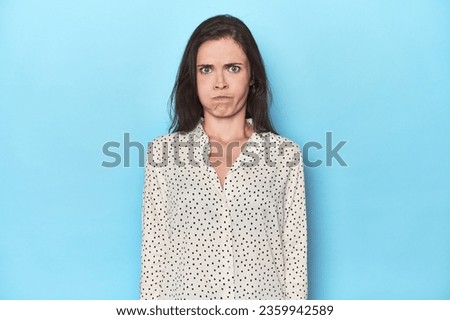 Young caucasian woman on blue backdrop blows cheeks, has tired expression. Facial expression concept.