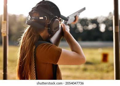 Young caucasian woman on tactical gun training classes. Woman with weapon, wearing cap, protective headphones and eyeglasses. Outdoor Shooting Range. At Sunny evening at summer. Rear view