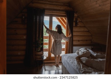 Young caucasian woman opening curtains in a wooden chalet cabin in the mountains. Comfortable rest in a log house in nature - Powered by Shutterstock