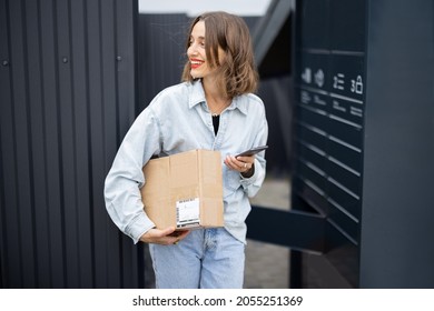 Young caucasian woman with parcel near automatic post terminal. Smiling girl holding smartphone and looking away on city street. Concept of smart delivery. Idea of modern shipping and logistics - Shutterstock ID 2055251369