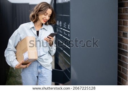Young caucasian woman with parcel near automatic post terminal. Smiling girl holding smartphone and looking away on city street. Concept of smart delivery. Idea of modern shipping and logistics