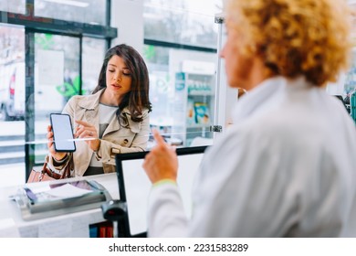 A young Caucasian woman is at the pharmacy showing her mobile phone to the pharmacist with a medication she is looking for. - Powered by Shutterstock