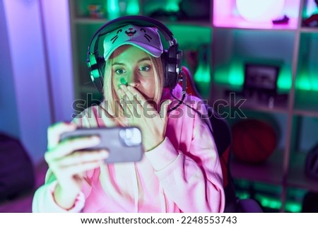 Young caucasian woman playing video games with smartphone scared and amazed with open mouth for surprise, disbelief face 