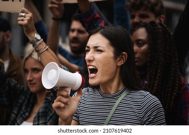 Young caucasian woman protester demonstrate shouting loud through megaphone while being on a protest - multicultural men and women protestors on a global strike demonstration - Shutterstock ID 2107619402