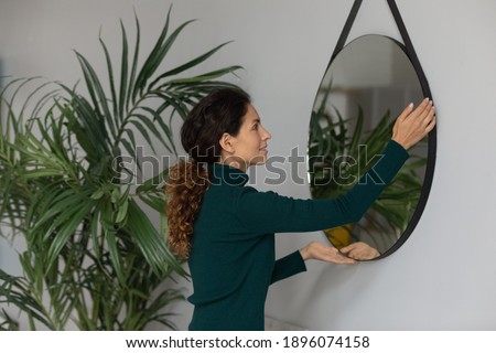 Young Caucasian woman put mirror on wall renovate modern living room in new own home. Millennial female renter or tenant decorate house or apartment moving in or relocating. Rent concept.