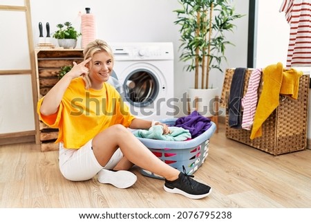 Young caucasian woman putting dirty laundry into washing machine smiling pointing to head with one finger, great idea or thought, good memory 