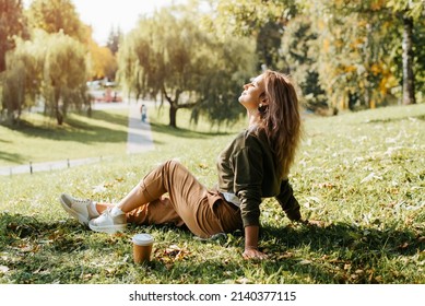 Young caucasian woman resting carefree, relaxing on lawn on sunny day. Side view hipster woman with closed eyes enjoying calmness and freedom during coffee break while sitting on grass outdoors.