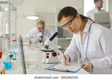 Young caucasian woman scientist in protective goggles making notes in report on clipboard sitting at desk with microscope. Female researcher working on vaccine development at biochemistry laboratory - Shutterstock ID 2022402101