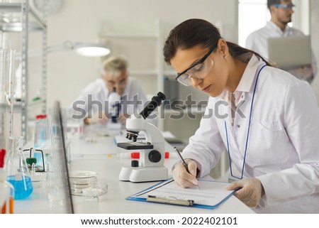 Young caucasian woman scientist in protective goggles making notes in report on clipboard sitting at desk with microscope. Female researcher working on vaccine development at biochemistry laboratory