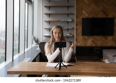Young Caucasian woman sit in home office record video live broadcast on smartphone camera. Millennial successful female coach or tutor talk on webcam zoom call on cellphone, have virtual event.