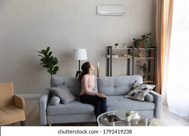 Young Caucasian woman sit on sofa in living room turn air condition device with remote controller, smiling millennial female relax on couch at home switch conditioner, breathe fresh air in apartment - Shutterstock ID 1792419238