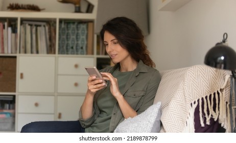 Young Caucasian woman sit rest on comfortable couch in living room look at cellphone screen shopping online on device. Happy millennial female relax on sofa at home text message on smartphone. - Shutterstock ID 2189878799