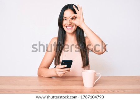 Young caucasian woman sitting at the desk using smartphone drinking coffee smiling happy doing ok sign with hand on eye looking through fingers 
