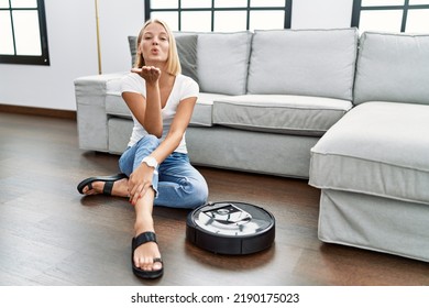 Young Caucasian Woman Sitting At Home By Vacuum Robot Looking At The Camera Blowing A Kiss With Hand On Air Being Lovely And Sexy. Love Expression. 