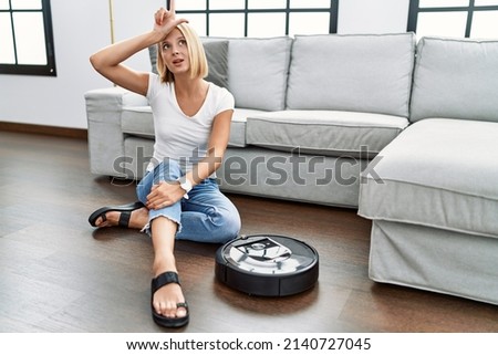 Young caucasian woman sitting at home by vacuum robot making fun of people with fingers on forehead doing loser gesture mocking and insulting. 