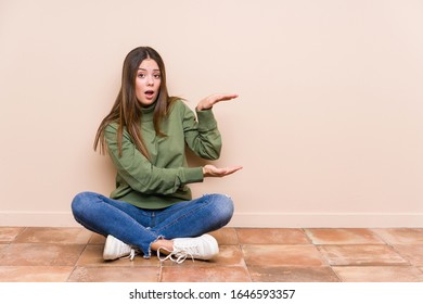 Young caucasian woman sitting on the floor isolated shocked and amazed holding a copy space between hands. - Shutterstock ID 1646593357