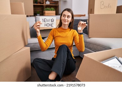 Young Caucasian Woman Sitting On The Floor At New Home Holding New Home Banner Smiling Looking To The Side And Staring Away Thinking. 