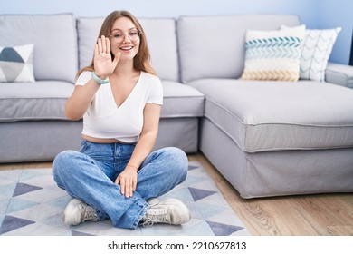 Young Caucasian Woman Sitting On The Floor At The Living Room Waiving Saying Hello Happy And Smiling, Friendly Welcome Gesture 