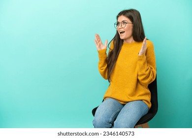 Young caucasian woman sitting on a chair isolated on blue background with surprise facial expression