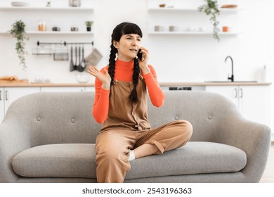 Young Caucasian woman sitting on sofa in modern kitchen having phone call. Wearing casual orange and brown outfit, she gestures expressively, engaged in conversation. - Powered by Shutterstock