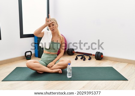Young caucasian woman sitting on training mat at the gym making fun of people with fingers on forehead doing loser gesture mocking and insulting. 