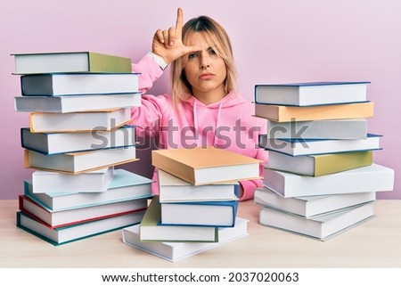 Young caucasian woman sitting on the table with books making fun of people with fingers on forehead doing loser gesture mocking and insulting. 