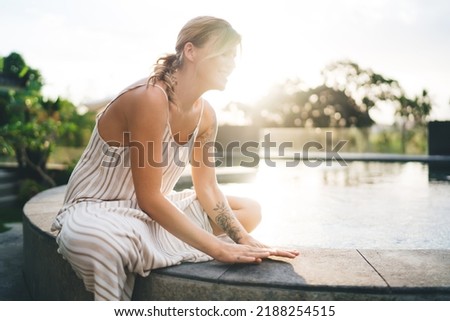 Young caucasian woman sitting and resting on swimming pool edge in yard of resort hotel. Tourism, vacation and weekend. Happy girl with tattoos. Sunset. Idyllic and tranquil lifestyle on Bali island