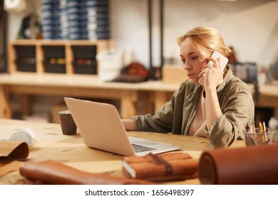 Young Caucasian Woman Sitting At Table In Leather Craft Workshop Talking On Phone And Using Laptop, Horizontal Portrait