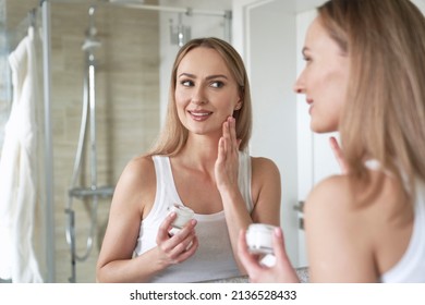 Young caucasian woman smiling and applying face cream in the mirror reflection