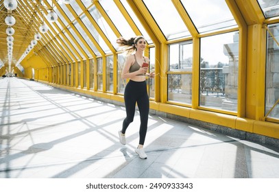 young Caucasian woman in sports bra and leggings jogs through yellow pedestrian bridge with smartphone and listening to music through earphones. energy and enthusiasm on sunny day during her workout - Powered by Shutterstock