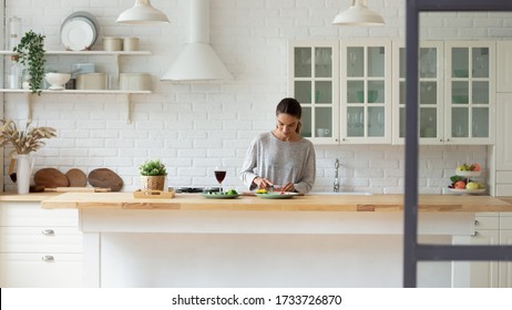 Young Caucasian woman stand at modern design kitchen counter cooking vegetable salad drinking wine, millennial girl female tenant or renter prepare vegetarian food in beautiful renovated home - Powered by Shutterstock