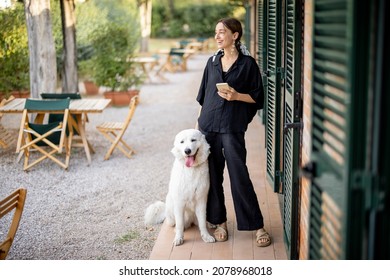 Young Caucasian Woman Standing With Maremmano-Abruzzese Sheepdog And Looking Away Near Entrance To Hotel Room. Concept Of Weekend, Rest And Vacation. Smiling Beautiful Girl With Smartphone