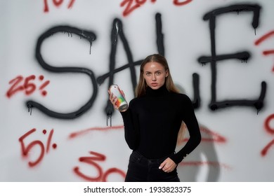 Young Caucasian Woman Standing Near Wall With Lettering Sale While Holding Paint Sprayer And Looking At Camera. Discount And Sale Concept