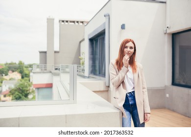 Young Caucasian Woman Standing On The Terrace Of A Modern Office Or Balcony At Home And Smokes Marijuana. Business Lady Smoking Cannabis. Female Relieves Stress At Work A Light Drug. Businesswoman