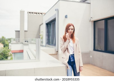 Young Caucasian Woman Standing On The Terrace Of A Modern Office Or Balcony At Home And Smokes Marijuana. Business Lady Smoking Cannabis. Female Relieves Stress At Work A Light Drug. Businesswoman