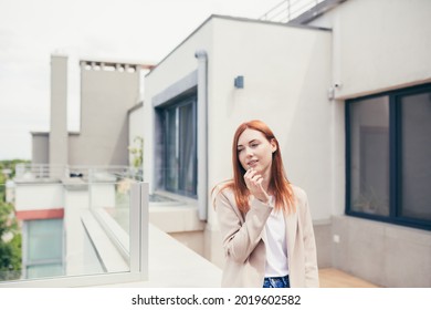 Young Caucasian Woman Standing On The Terrace Of A Modern Office Or Balcony At Home And Smokes Marijuana. Business Lady Smoking Cannabis. Female Relieves Stress At Work A Light Drug. Businesswoman