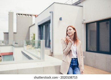 Young Caucasian Woman Standing On The Terrace Of A Modern Office Or Balcony At Home And Smokes Marijuana. Business Lady Smoking Cannabis. Female Relieves Stress At Work A Light Drug. Businesswoman