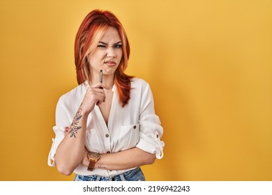 Young Caucasian Woman Standing Over Yellow Background Holding Pen Clueless And Confused Expression. Doubt Concept. 