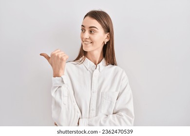Young caucasian woman standing over isolated background smiling with happy face looking and pointing to the side with thumb up.  - Powered by Shutterstock