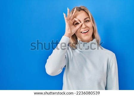 Young caucasian woman standing over blue background doing ok gesture with hand smiling, eye looking through fingers with happy face. 