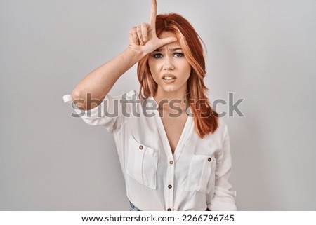 Young caucasian woman standing over isolated background making fun of people with fingers on forehead doing loser gesture mocking and insulting. 