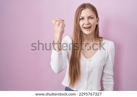 Young caucasian woman standing over pink background angry and mad raising fist frustrated and furious while shouting with anger. rage and aggressive concept. 