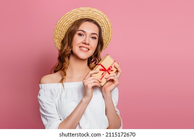 Young caucasian woman in straw hat and summer clothes holding small paper craft box with red ribbon and smiling isolated over pink background. Holiday greeting, surprise. copy pace