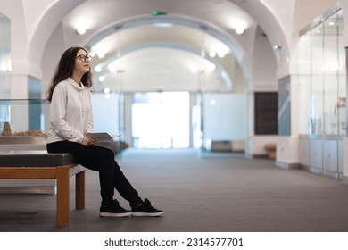 Young Caucasian woman student sitting on bench in museum visiting educational excursion. Light hall in background. Concept of cultural exhibition. - Powered by Shutterstock