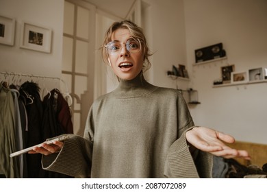 Young Caucasian Woman With Surprised Face Waving Hands In Shock From News She Read On Phone. Blonde With Her Hair Pulled Back Wears Glasses And Sweater. Emotion Concept, Facial Expression.