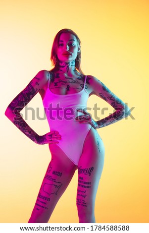 Young caucasian woman in swimsuit posing stylish on gradient yellow studio background in neon light. Beautiful model with tattoos. Human emotions, sales, ad concept. Resort and vacation, summertime.