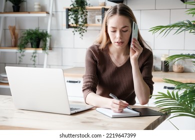 Young Caucasian Woman Talking With Client On Smartphone And Writing In Notepad While Sitting At The Desk And Working On Remote Project At Home